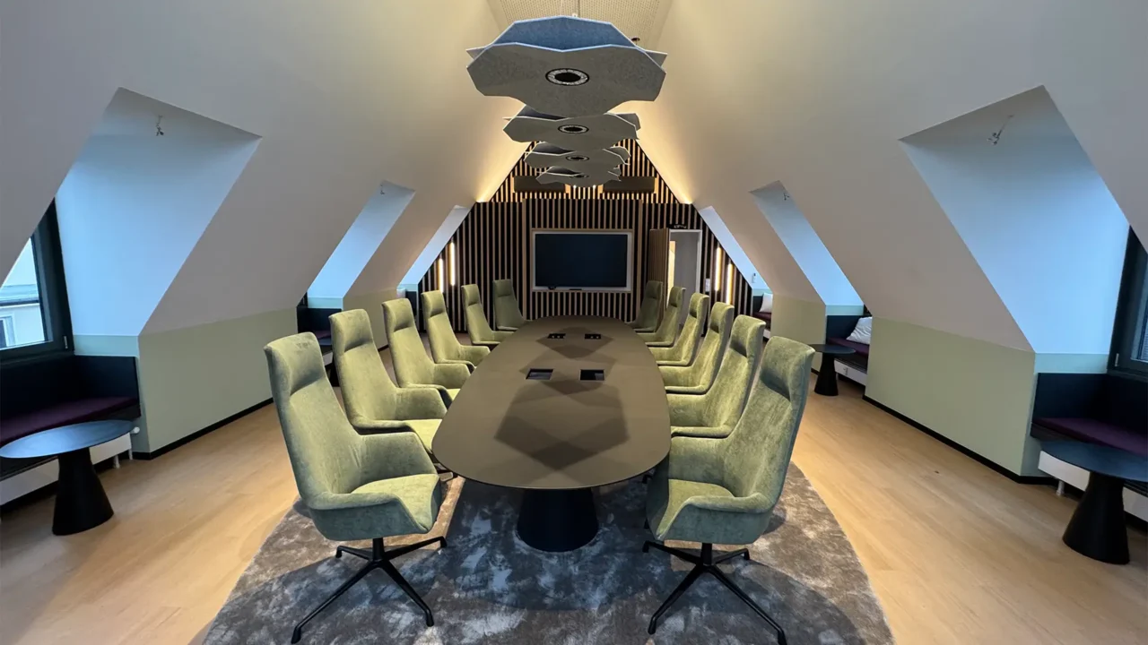 Großer Konferenzraum mit langem Tisch, grünen Polsterstühlen, Akustikelementen an der Decke und Bildschirm an der Stirnwand.