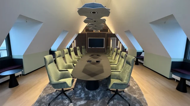 Großer Konferenzraum mit langem Tisch, grünen Polsterstühlen, Akustikelementen an der Decke und Bildschirm an der Stirnwand.