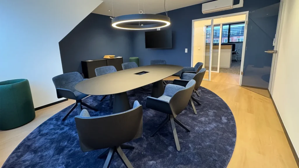Moderner Besprechungsraum mit ovalem Tisch, gepolsterten Stühlen, blauem Teppich und ringförmiger Deckenleuchte.
